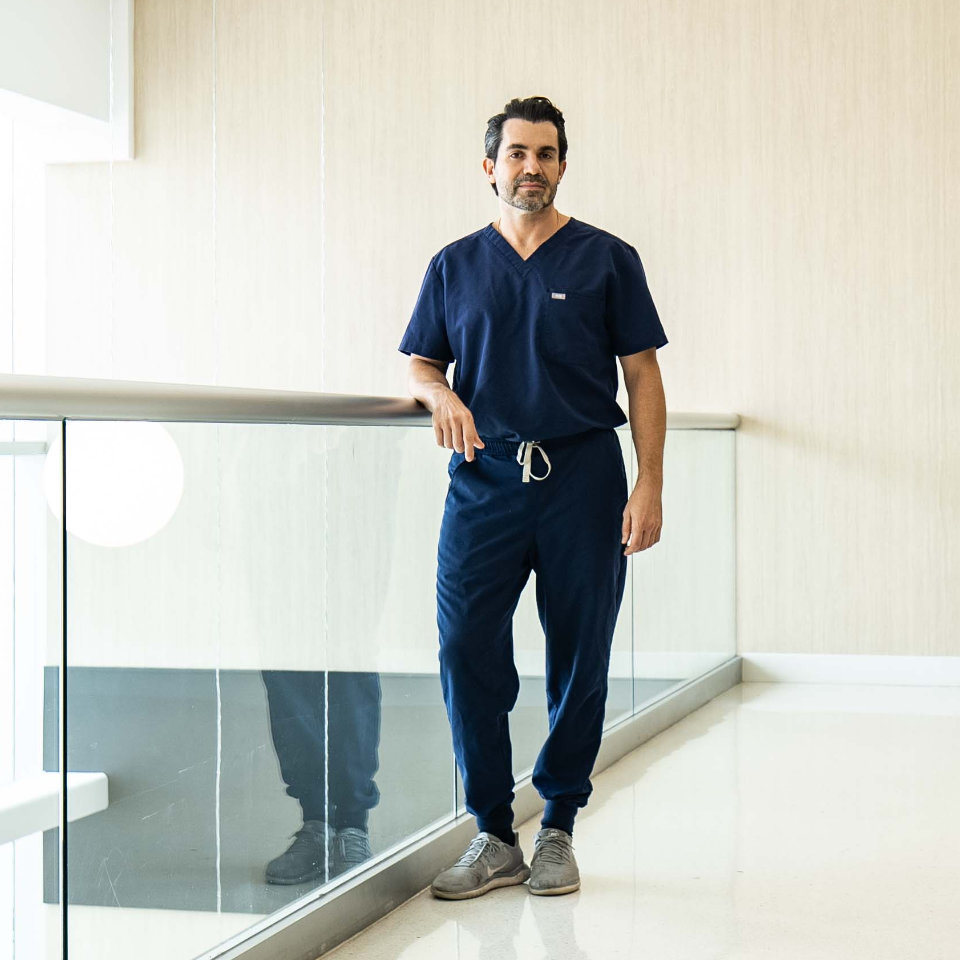 A man in navy blue scrubs stands by a glass railing in a modern, well-lit hallway with white floors and beige walls.