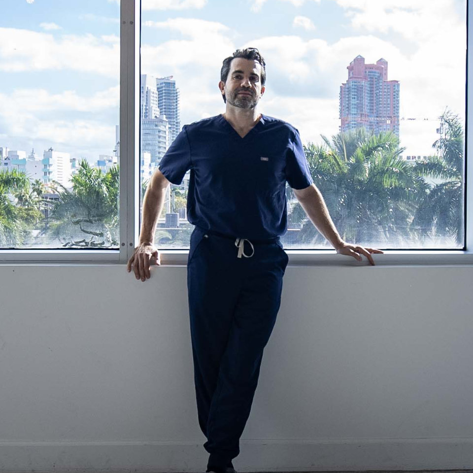 Un hombre con un uniforme azul marino está de pie junto a una gran ventana con el horizonte de la ciudad, palmeras y cielo azul visibles en el exterior.