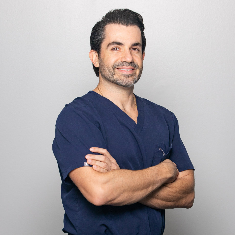 Un hombre con cabello oscuro y barba, que viste un uniforme médico azul marino, está de pie contra un fondo gris claro liso con los brazos cruzados y sonriendo.