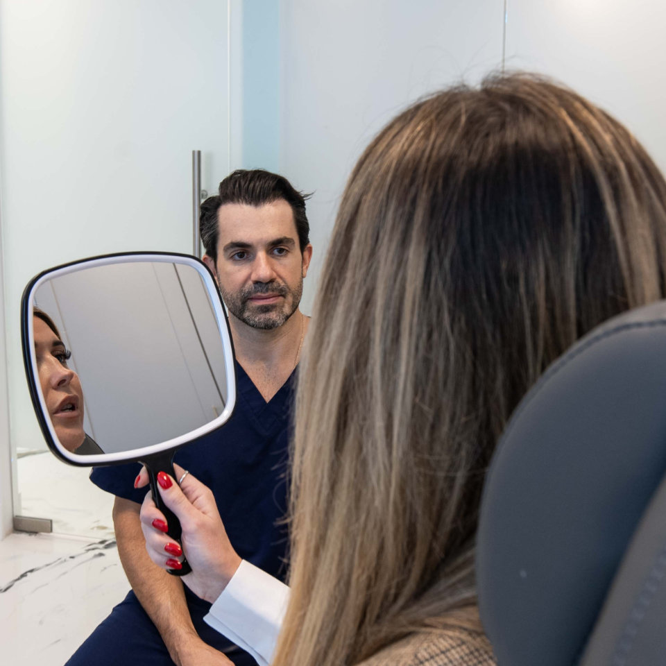 A woman holds a mirror in front of her face while sitting in a chair, with a man in medical scrubs seated across from her in a consultation room.