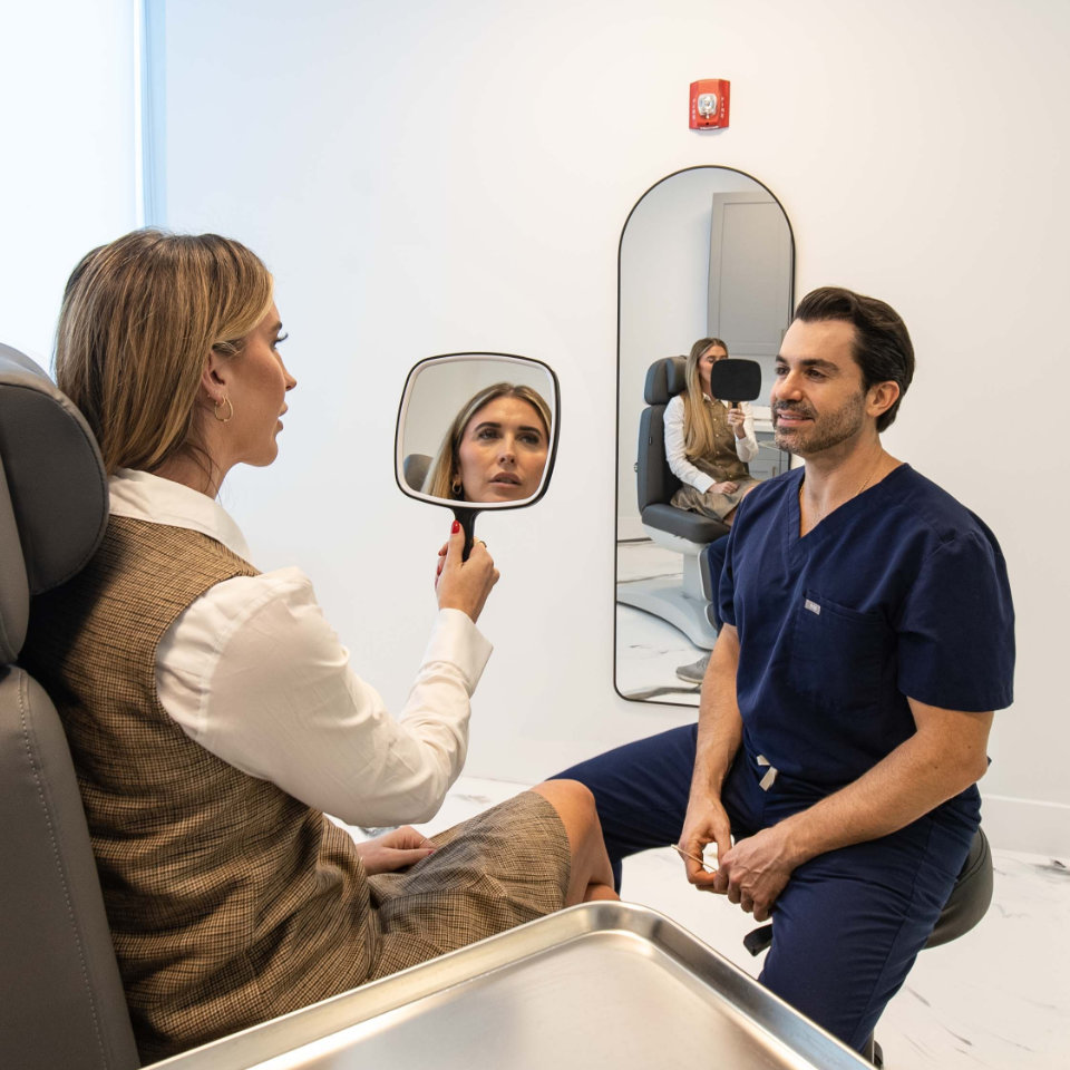 A woman examines her reflection with a handheld mirror while seated across from a man in medical scrubs in a modern clinic room.