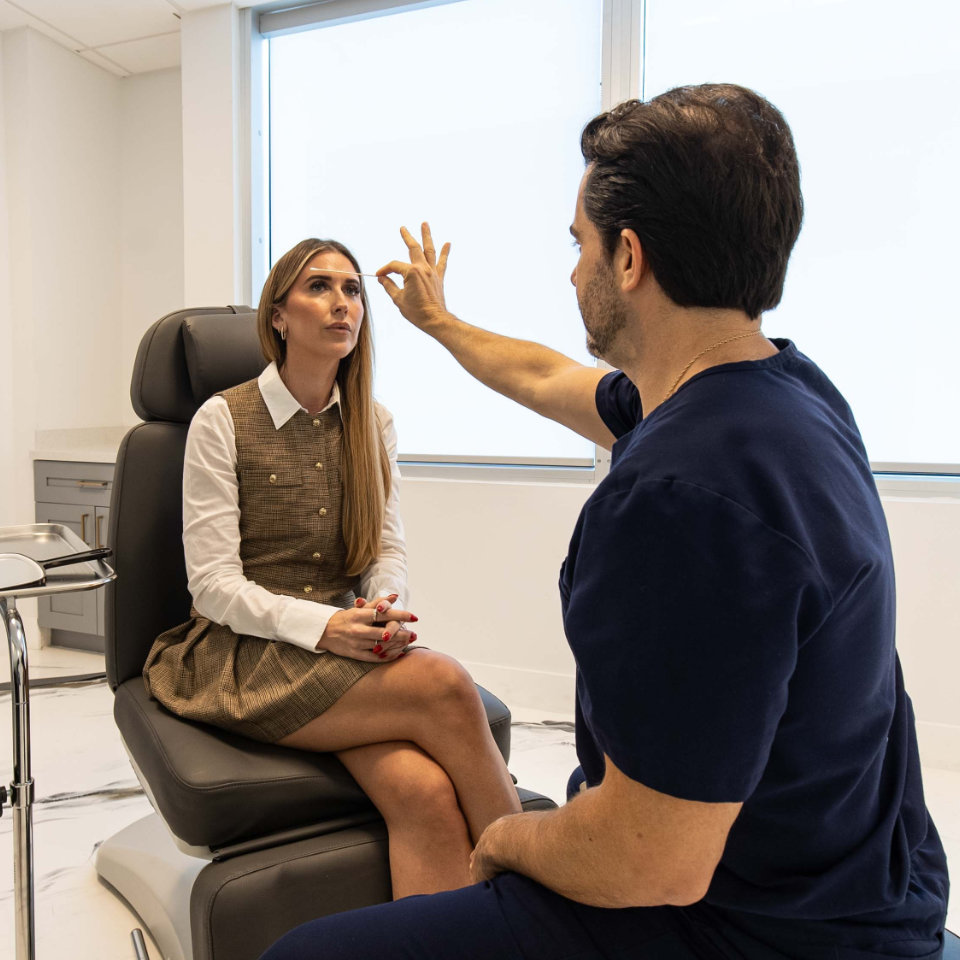 A woman sits in a medical chair while a healthcare professional in scrubs examines her face in a well-lit clinic room.