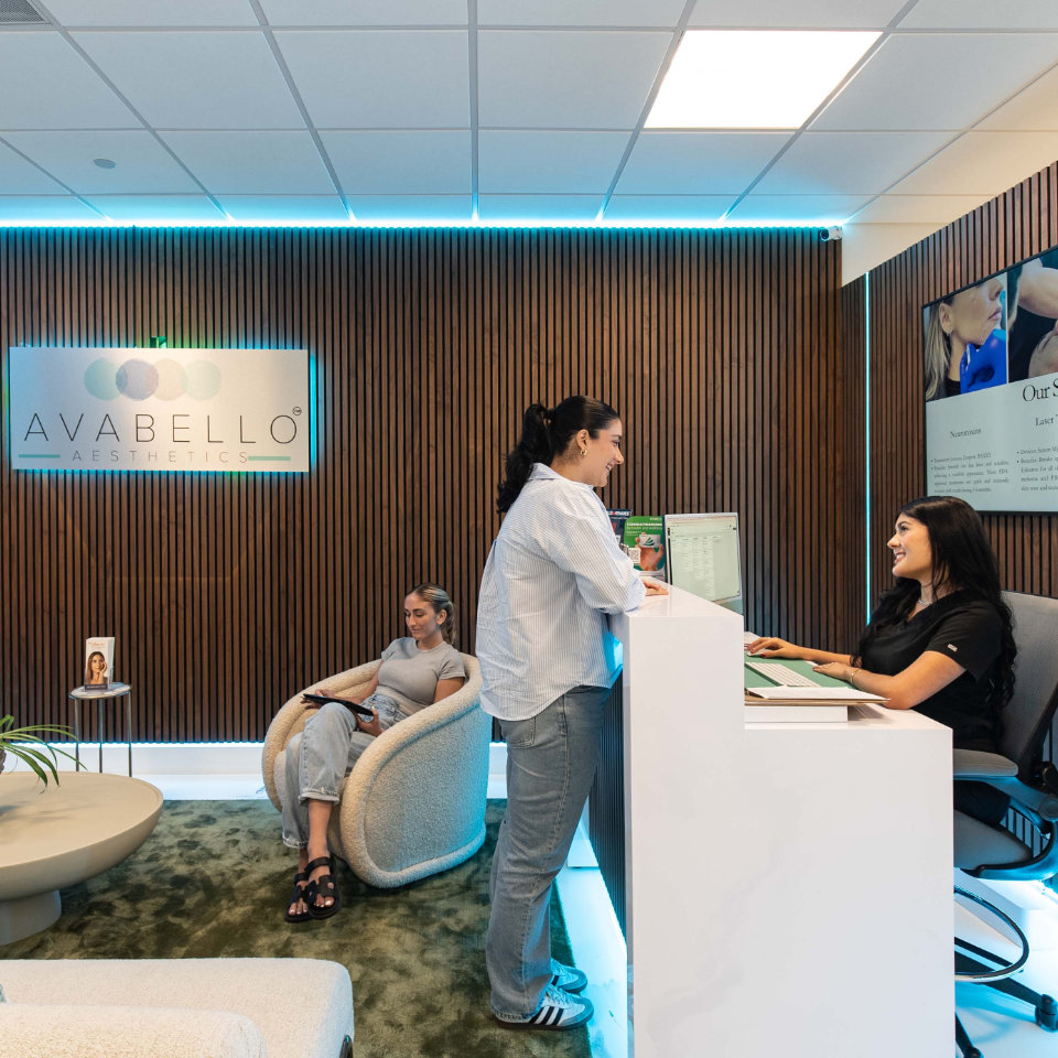 A receptionist assists a woman at the front desk of a modern aesthetics clinic, while another woman sits in the waiting area.