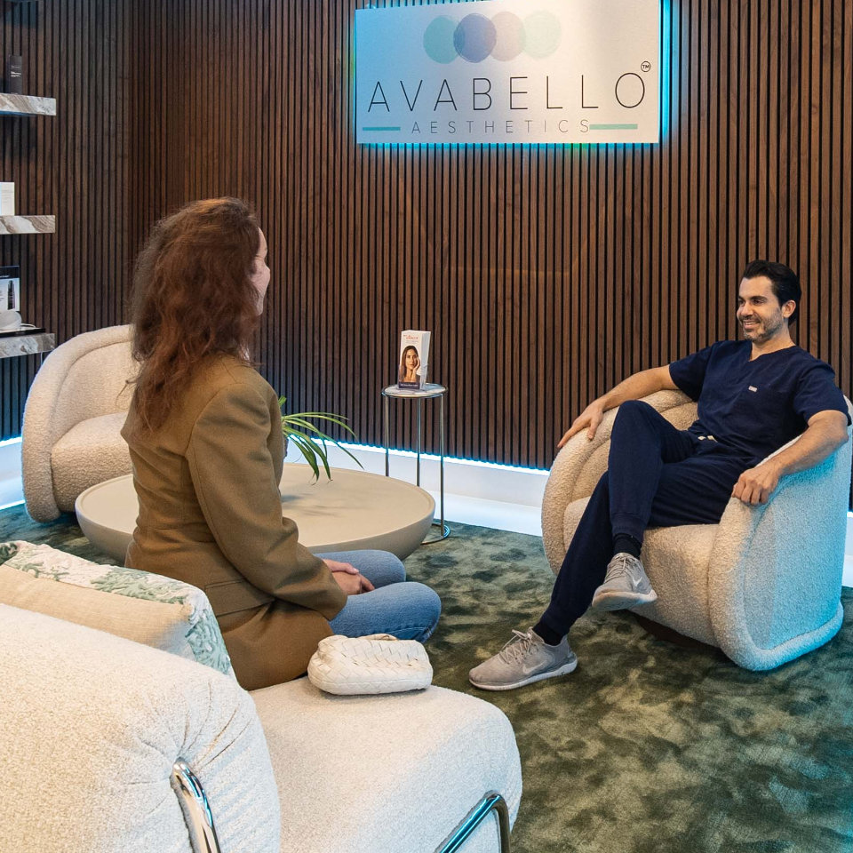 A woman and a man sit across from each other in a modern, softly lit aesthetic clinic waiting area with the sign "AVABELLO AESTHETICS" on the wall.