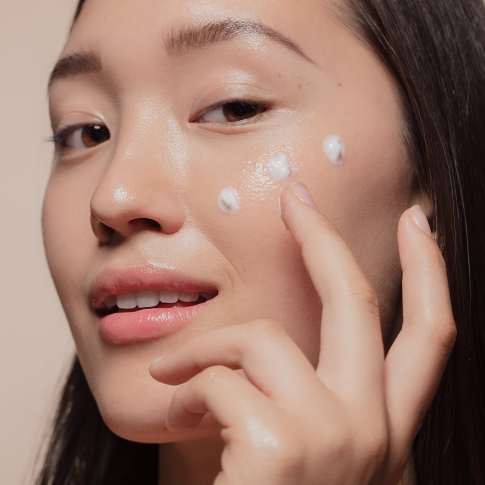 A woman applies small dots of cream to her cheek with her fingers, showcasing a skincare routine.